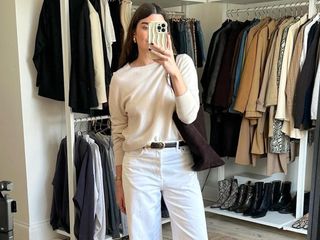 British influencer Anna Newton poses for a mirror selfie in her closet wearing a beige lightweight crewneck sweater, brown suede tote bag, black belt, and white jeans.