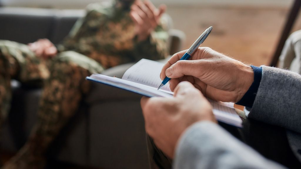 therapist taking notes, man in military uniform sits on couch behind