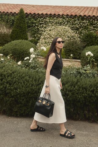 a photo showing what shoes to wear with skirts with a woman standing in a garden wearing a black waistcoat with a white midi skirt and black flat sandals