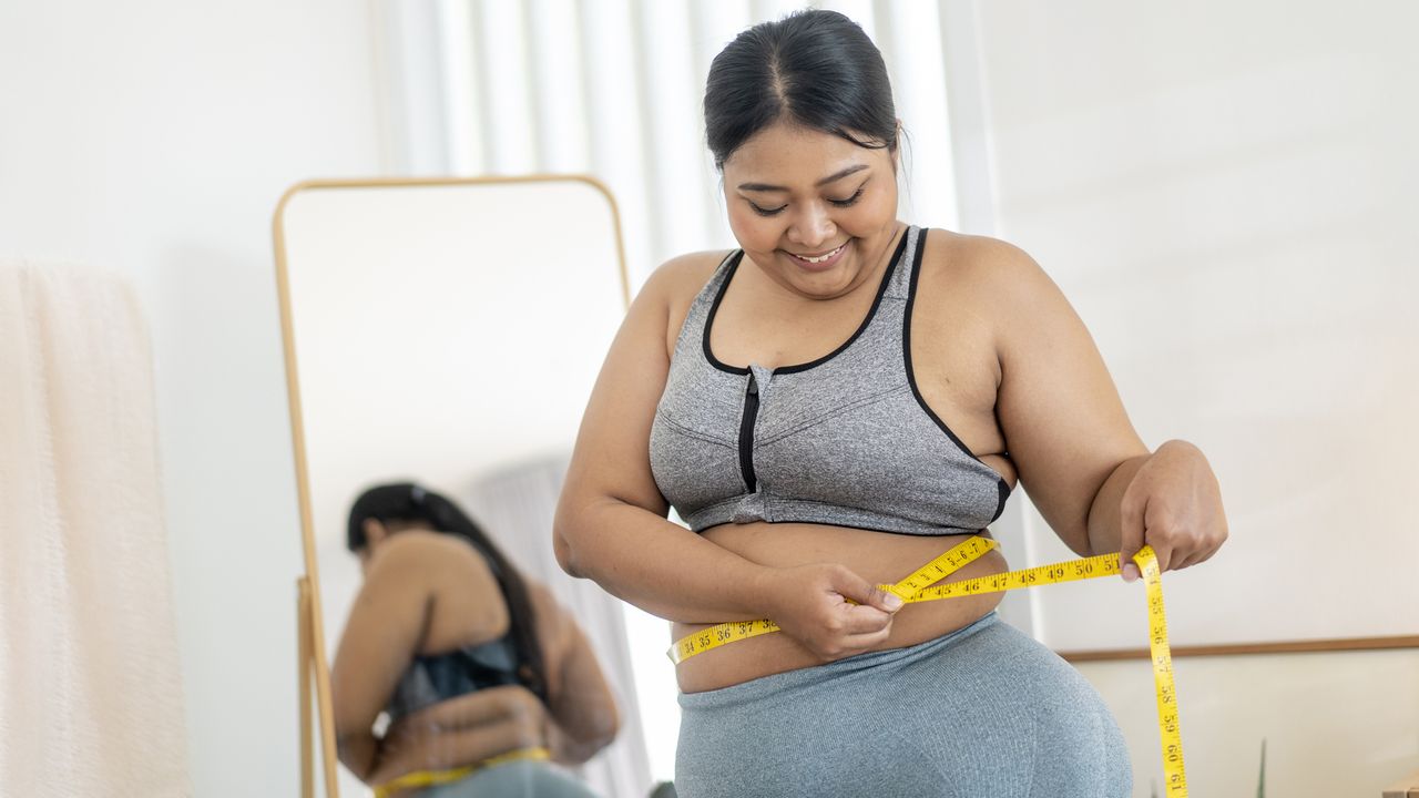 Smiling woman measuring her waist with a tape measure in bedroom