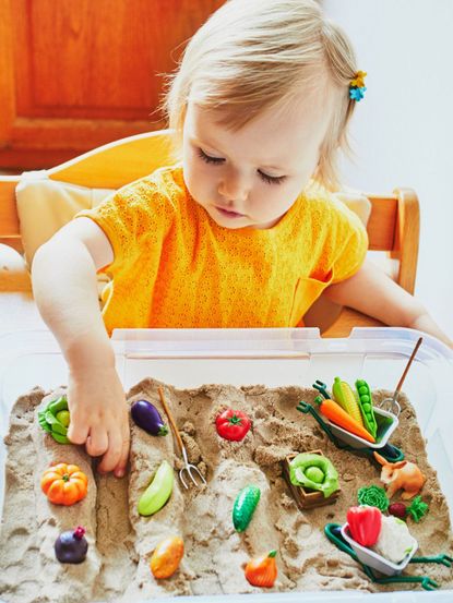 Child Playing With A Container Full Of Sand And Plastic Vegetables