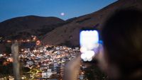 a person hold up a phone to take a photo of a total lunar eclipse occurring above a hills with city lights below.