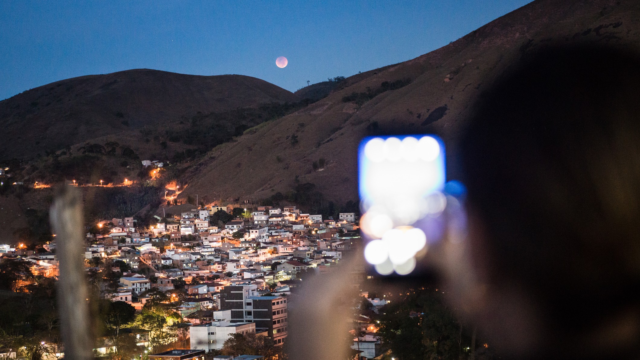 a person hold up a phone to take a photo of a total lunar eclipse occurring above a hills with city lights below.