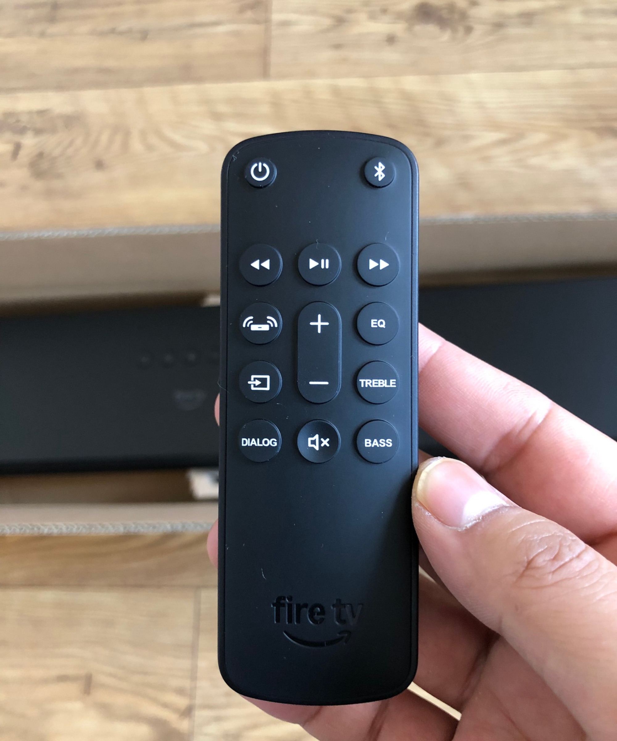 Hand holding the remote for the Fire TV Soundbar Plus with soundbar and wooden floor in background, in Dan Fauzi's home.