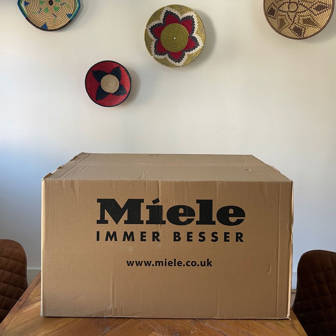 Brown box of the Miele Guard M1 Cat &amp; Dog sitting on a table in front of a white wall with colourful baskets on it
