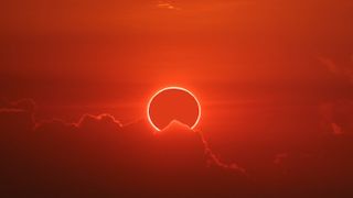 Solar eclipse photographed in Chon Buri, Thailand. The red hue image shows a distinct 'ring of fire' glowing in the sky, slightly obscured by a cloud. 