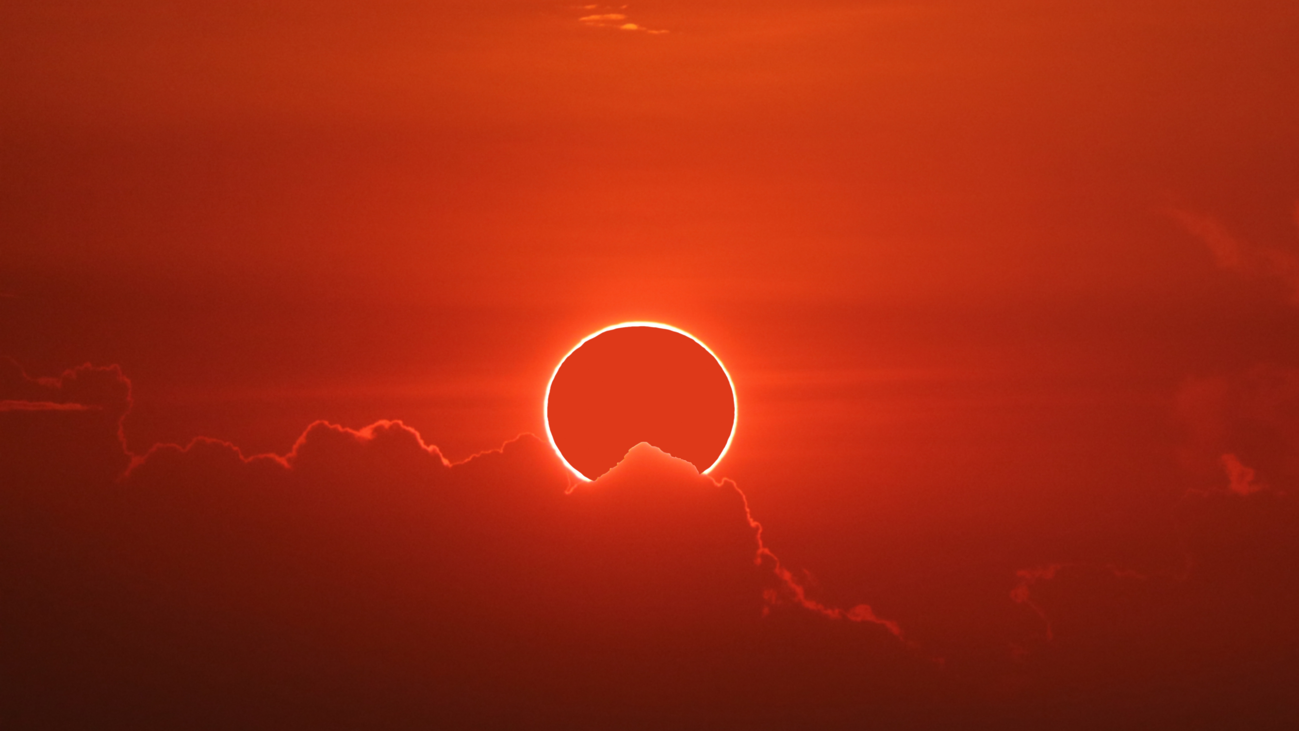 Solar eclipse photographed in Chon Buri, Thailand. The red hue image shows a distinct 'ring of fire' glowing in the sky, slightly obscured by a cloud.