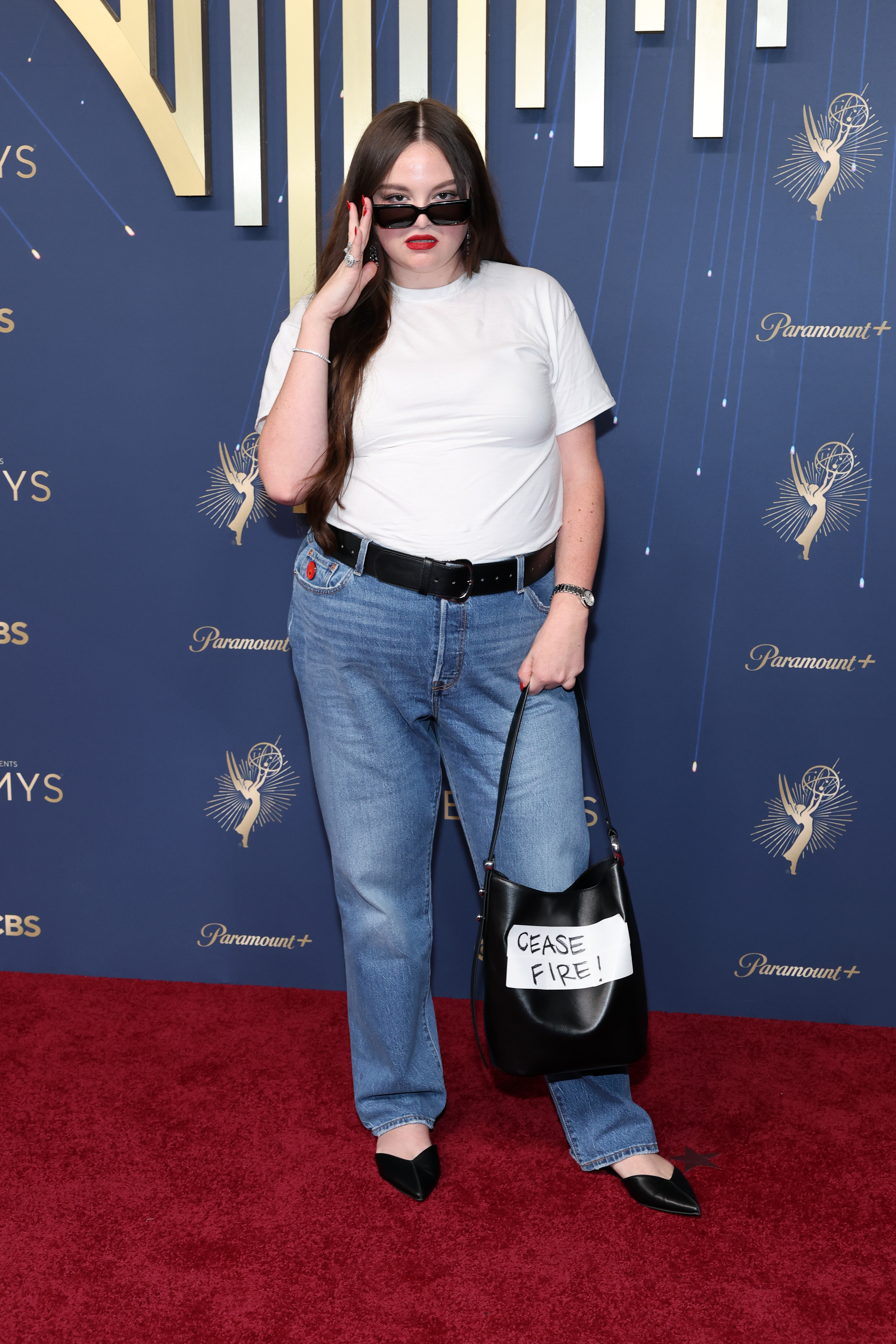Meg Stalter wearing a white T-shirt and jeans on the attends the 2025 Emmy Awards red carpet with a bag that reads Cease Fire!