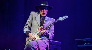 Steve Vai is suited and booted in grey and takes a solo on his Ibanez PIA onstage with Beat