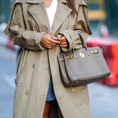 Tamara Kalinic wears quiet luxury nails, white wavy neck top, a beige long trench coat, a blue denim short skirt, a gray taupe Birkin handbag from Hermes