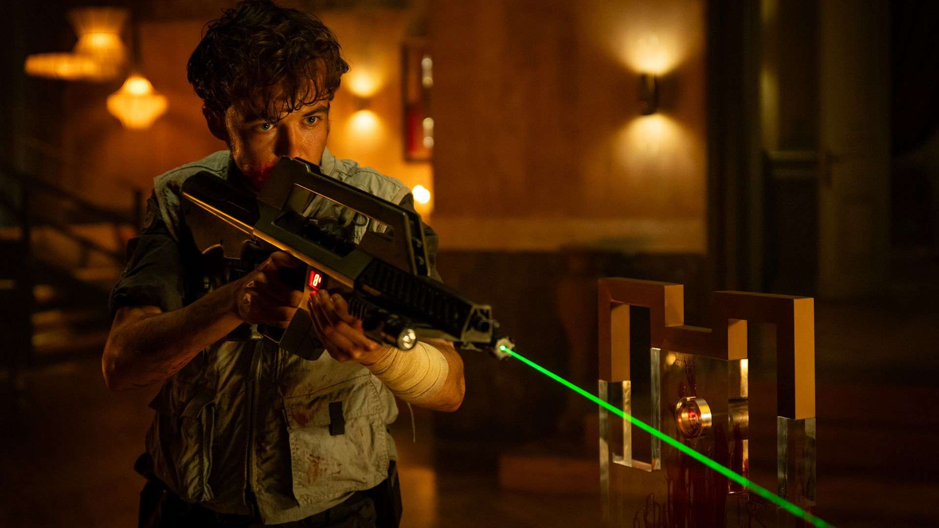 Screenshot from Alien: Earth. A young man holding a sci-fi assault rifle with a green laser sight affixed to the front. He&#039;s in an ornate room, with blood splattered around.