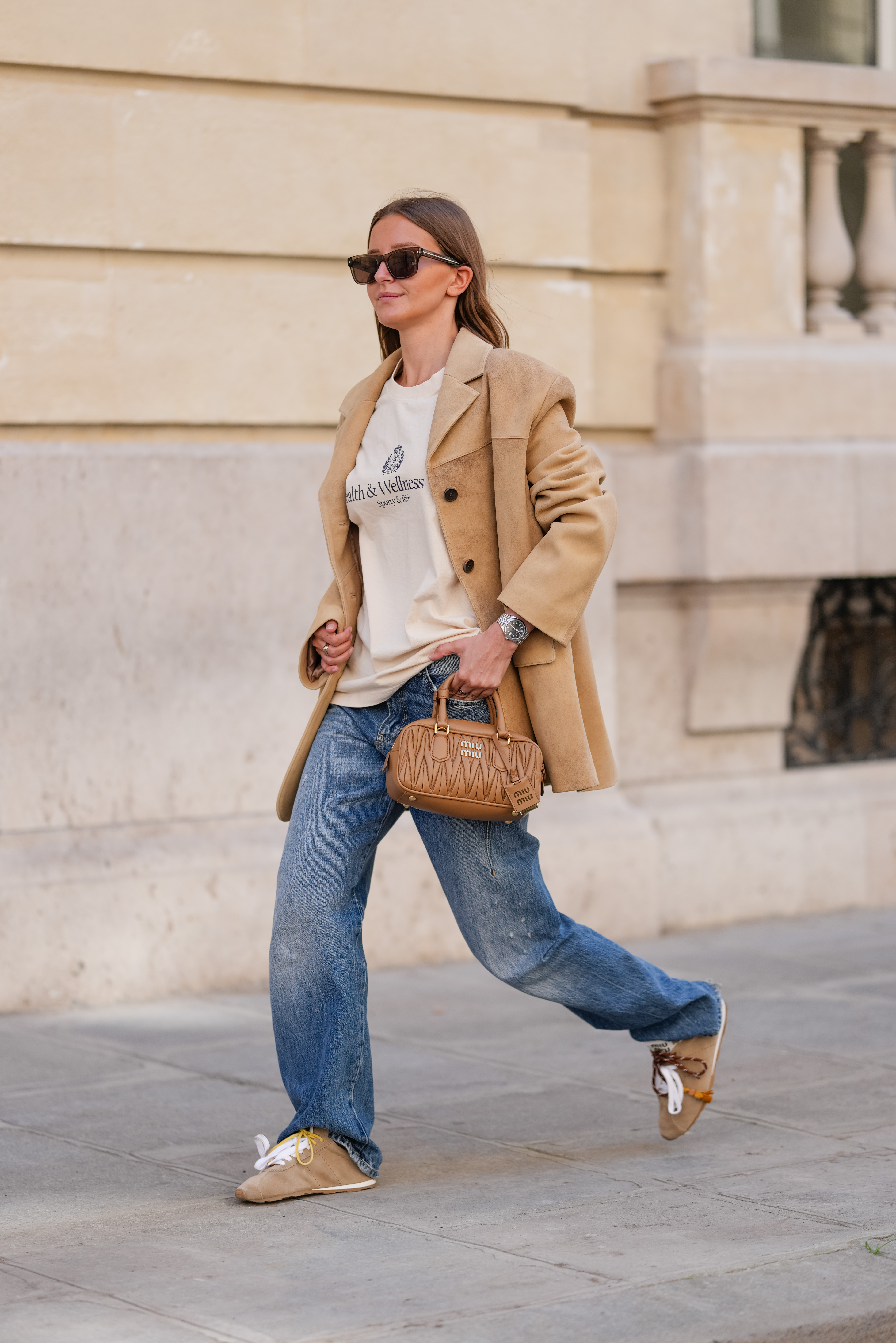 PARIS, FRANCE - APRIL 28: Diane Batoukina wears a beige Sporty & Rich logo T-shirt tucked into blue straight-leg Zara jeans, layered under a camel-toned oversized blazer jacket by Source Unknown. A pair of tan Miu Miu sneakers with mismatched laces in yellow and brown are worn with the cuffs of the jeans rolled. A quilted brown leather Miu Miu handbag with gold hardware is held by hand. Rectangular black Prada sunglasses with reflective lenses complete the look. Full body shot, during a street style fashion photo session, on April 28, 2025 in Paris, France. (Photo by Edward Berthelot/Getty Images)