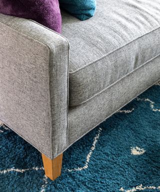 Corner of gray couch with wood legs on blue rug with white detail