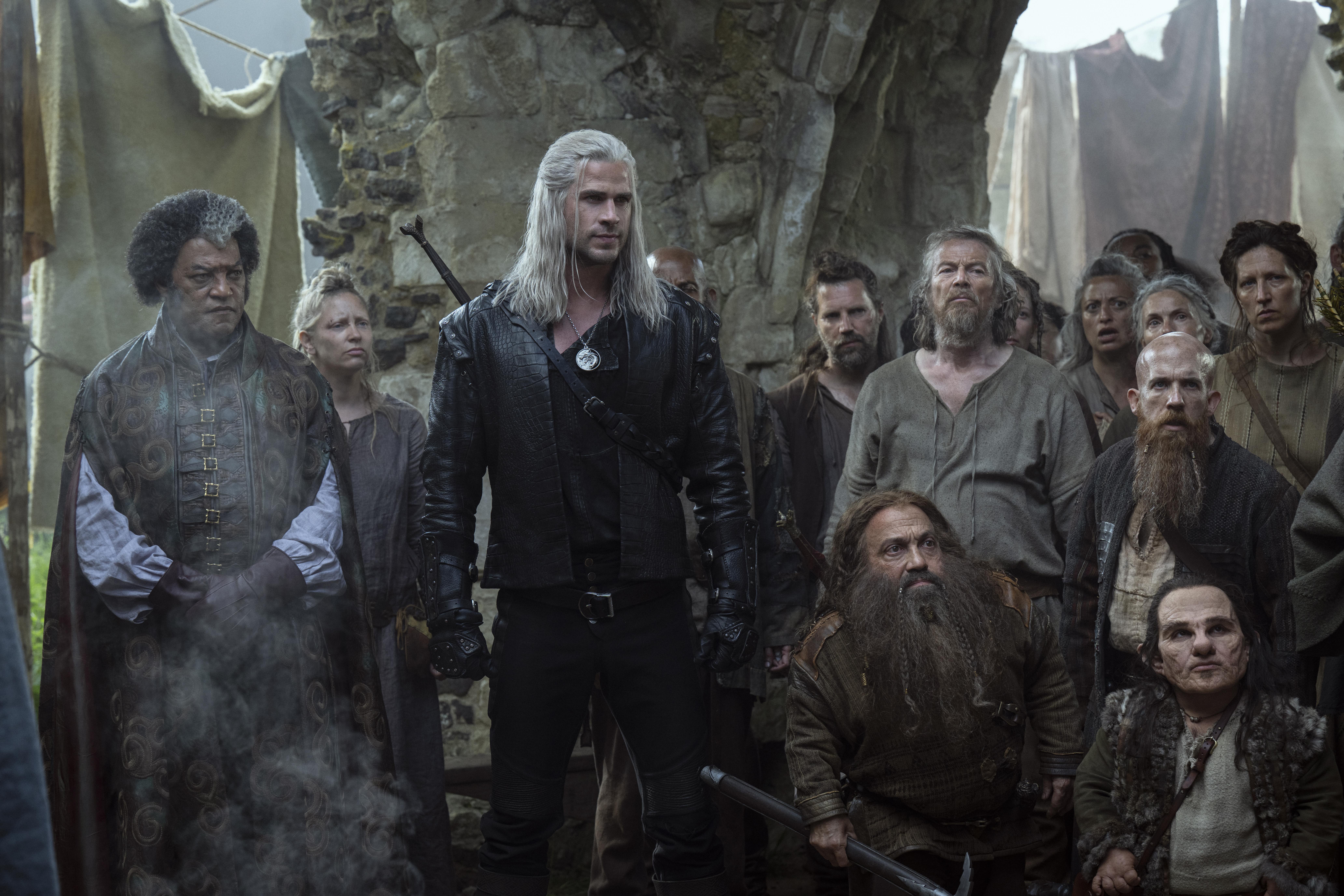 laurence fishburne and liam hemsworth surrounded by a crowd of people in medieval clothing in a still from the witcher season 4