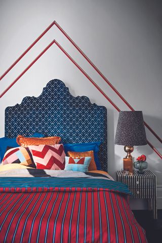Double bed with a blue patterned bedhead, with mixed patterned cushions and striped sidetable and bed covers in red and blue.