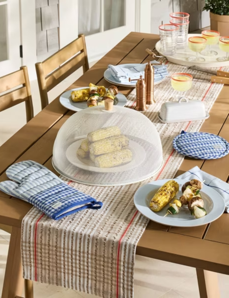 Outdoor tablescape featuring striped and gingham table linens, melamine plates, and a stack of corn on the cob, concealed with a metal food cover