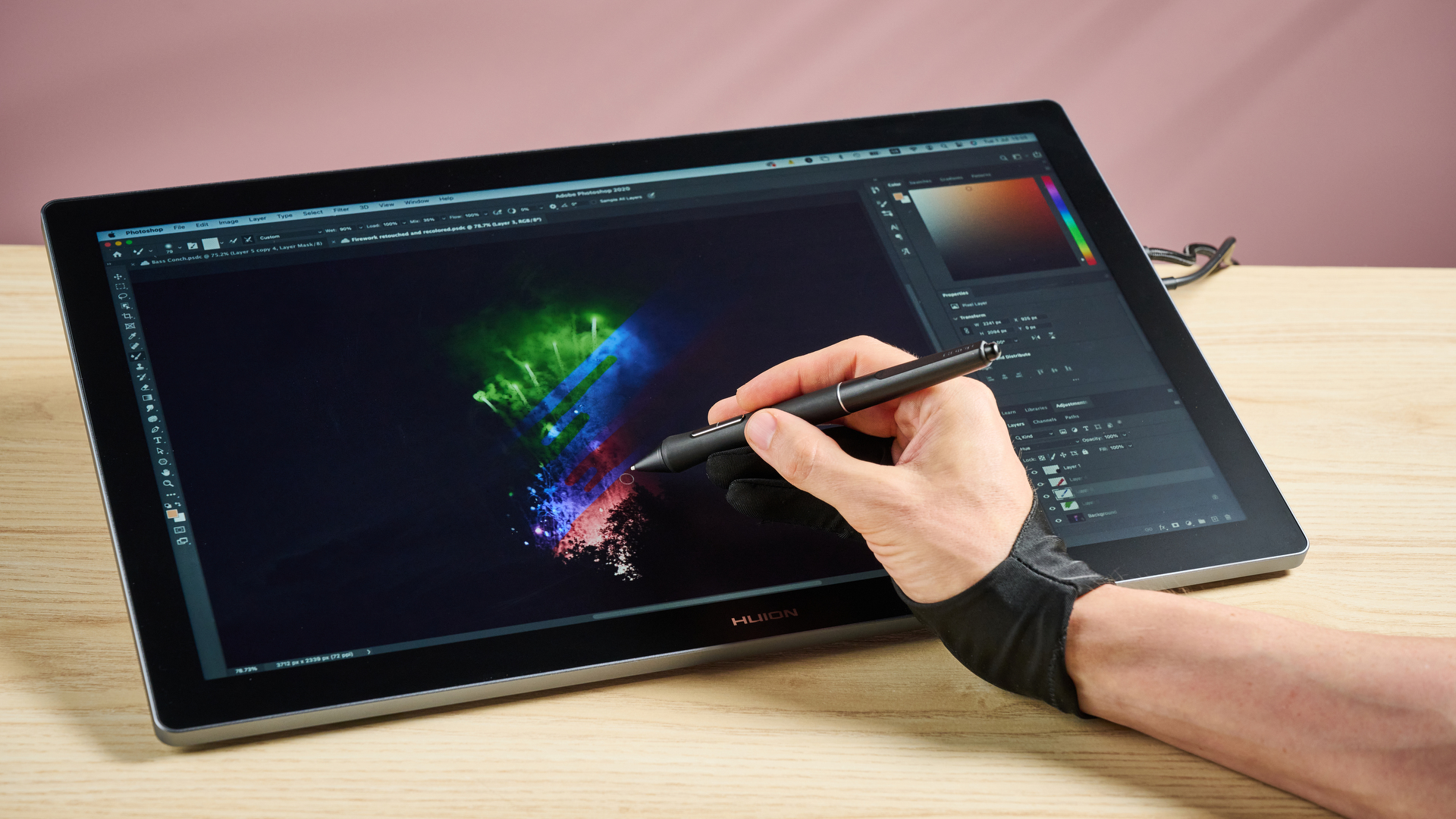 A man's hand holding a stylus and recoloring an image of fireworks on the Huion Kamvas Pro 19, in front of a pink background.