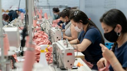photo of women wearing face masks as they sit at a long table and work with pink fabric at sewing machines 