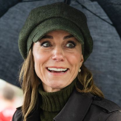 Princess Kate wearing a green cap holding an umbrella and grimacing