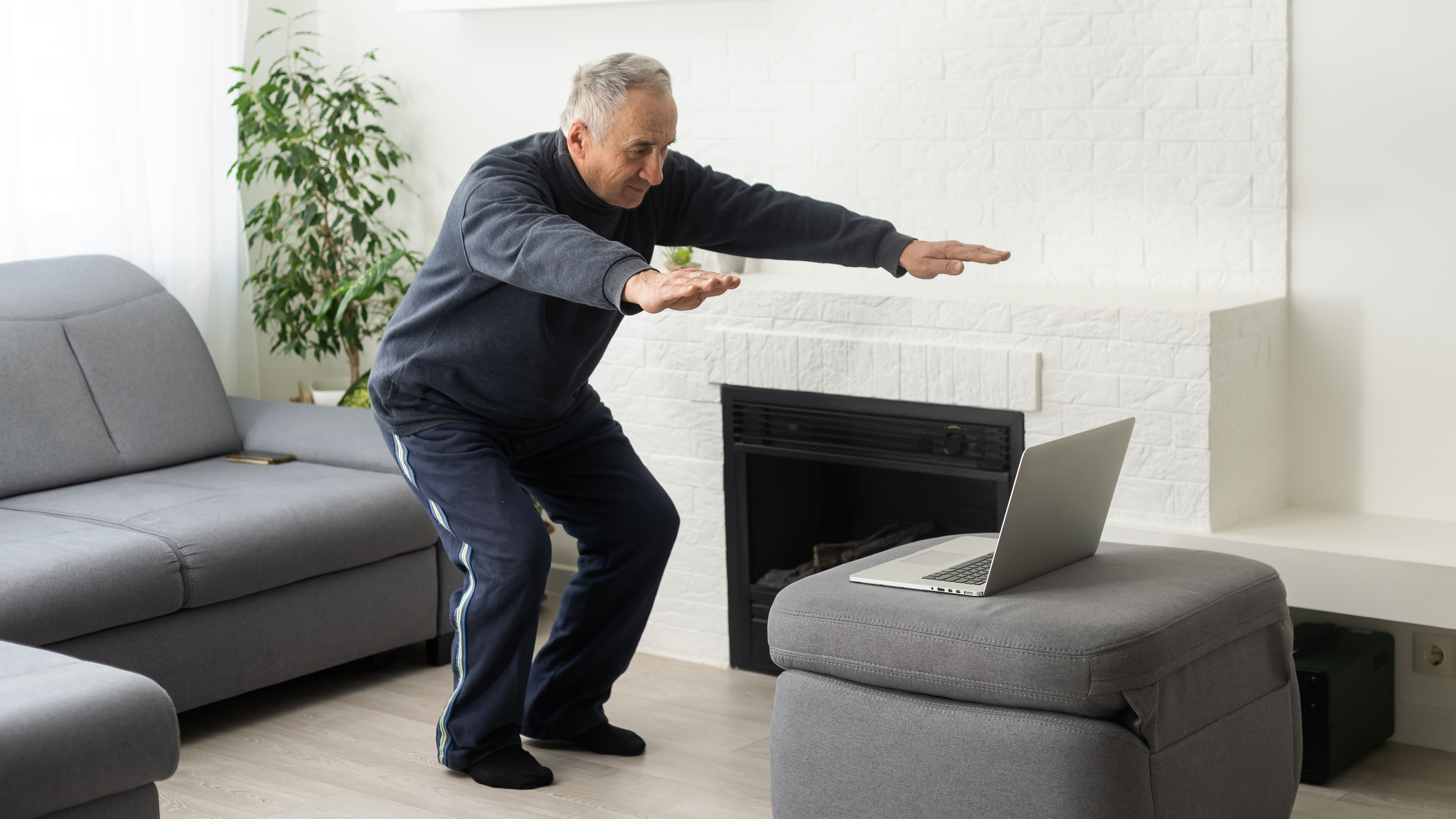 A man performs a shallow squat with his arms outstretched in his living room. Behind him is a couch and there's a laptop on a footstool in front of him.