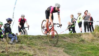 A rider on a red bike in a black and pink jersey nearly airborne over a grass jump