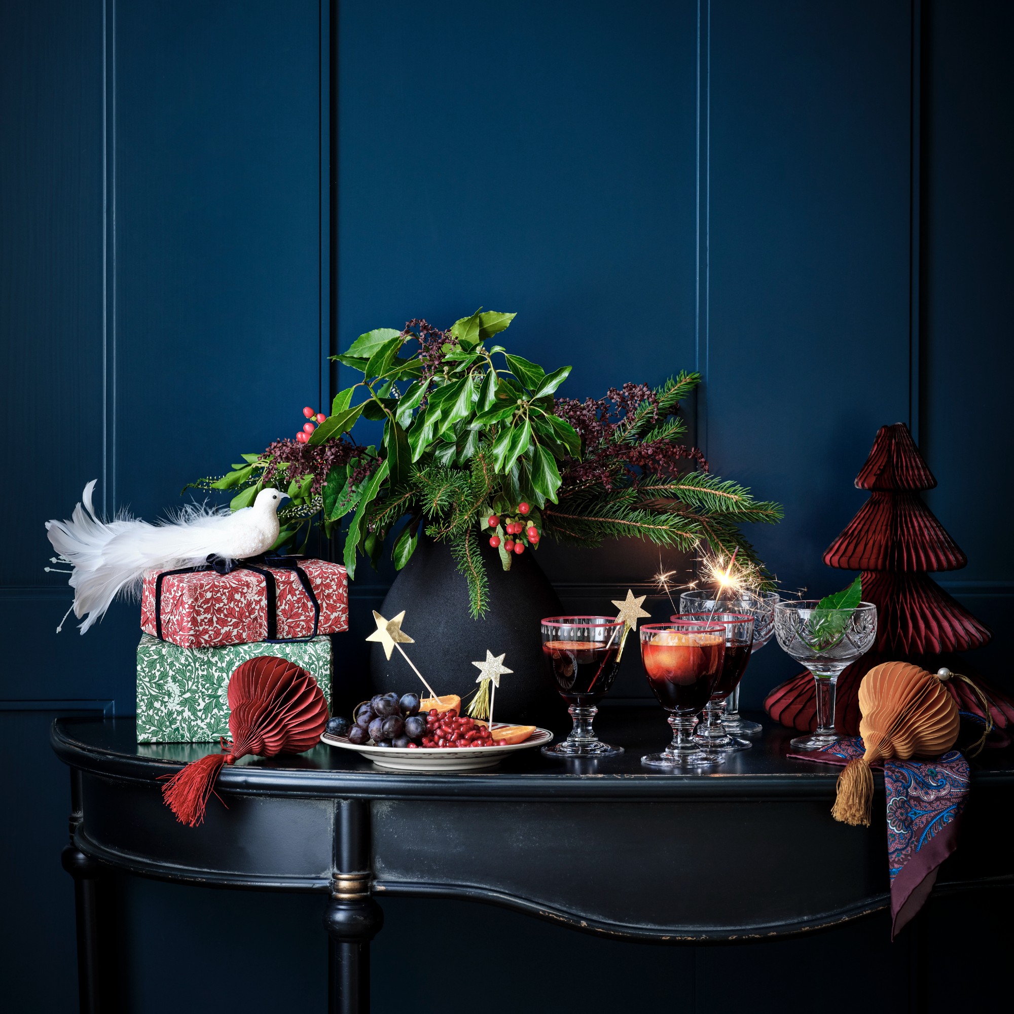 A console table against a navy panelled wall adorned with tasselled Christmas decorations, Christmas presents and a centrepiece