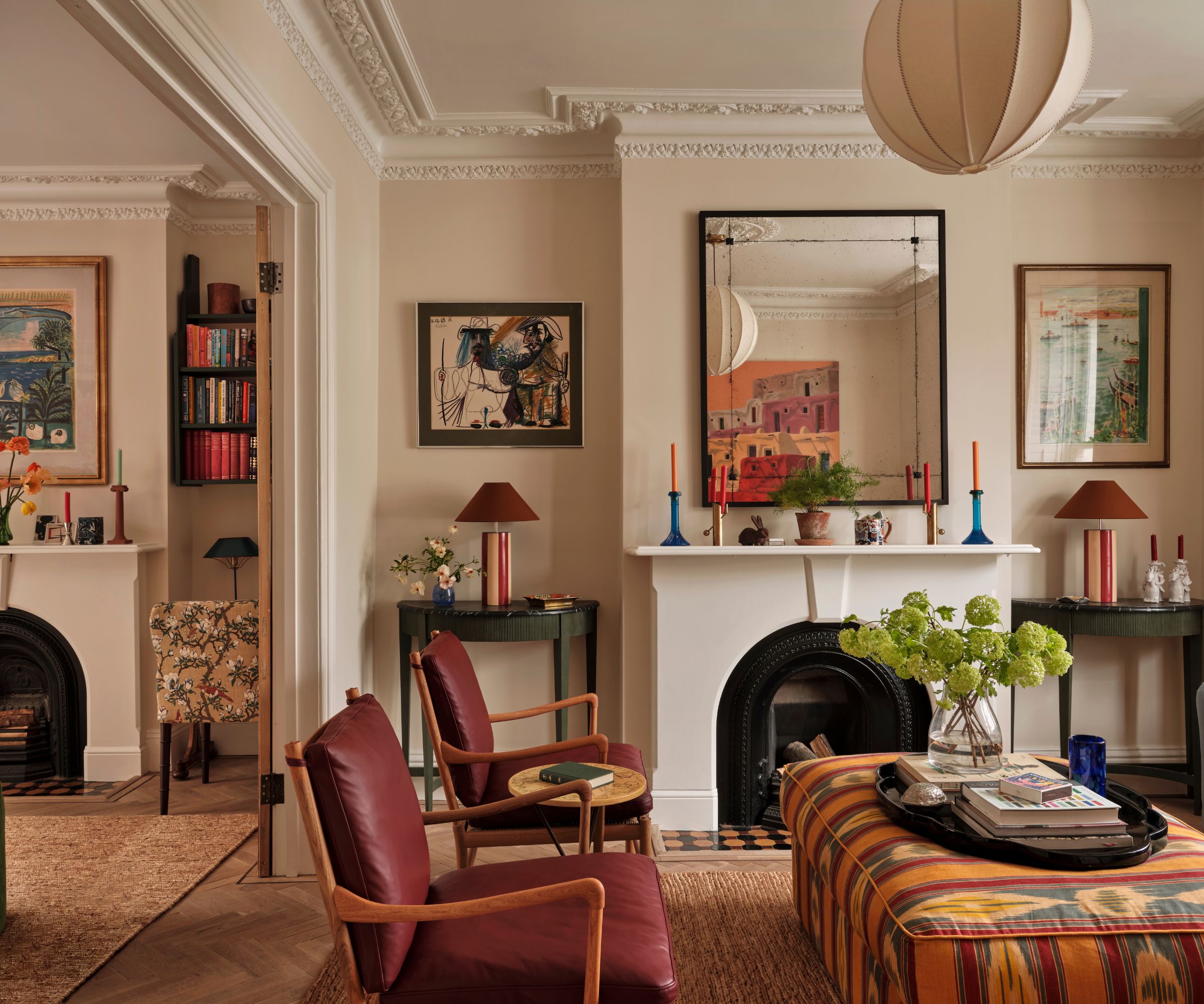 Eclectic living room with cream walls and white crown moulding, fireplace with a mirror above, lantern pendant light, two red leather lounge chairs, a colorful ottoman, and a vase of green flowers.