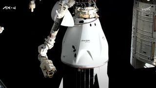 A white space capsule hangs upright, docked to the port of a space station. A robotic arm hangs to the left in the foreground.