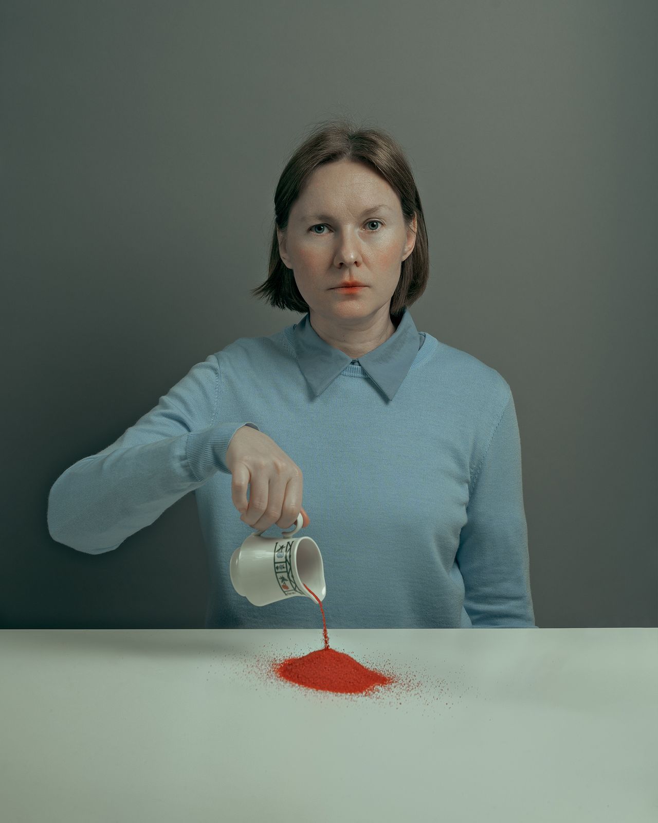 A woman in a light blue sweater pours red powder from a small container onto a white table, having red powder on her mouth too