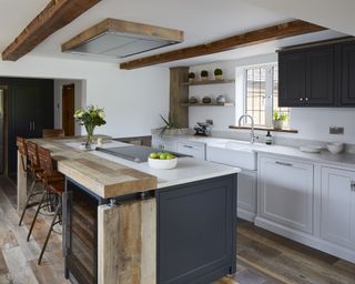 A kitchen with blue-grey island with reclaimed wood-topped breakfast bar, exposed wooden beams on a white ceiling, and white quartz worktops over pale grey cabinets