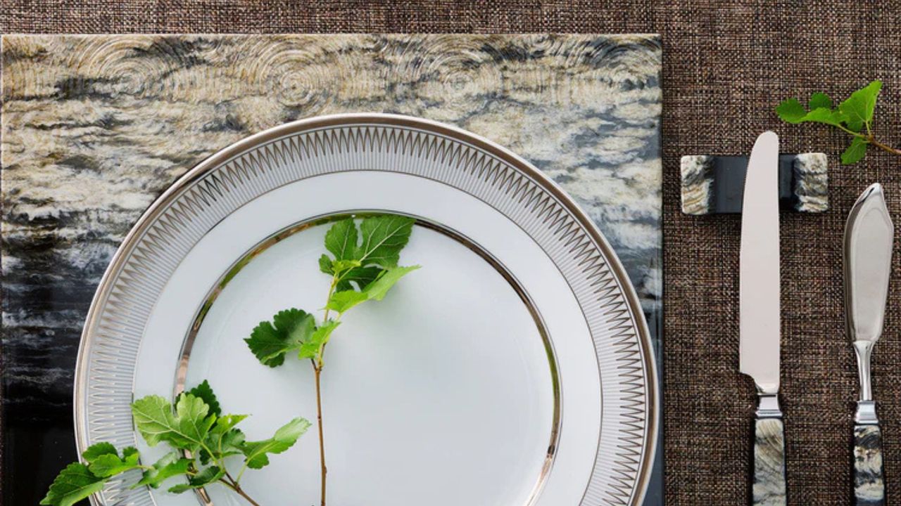 table setting with two stacked plates with foliage on it, a placemat, a knife resting on a cutlery rest, and another bread knife beside it