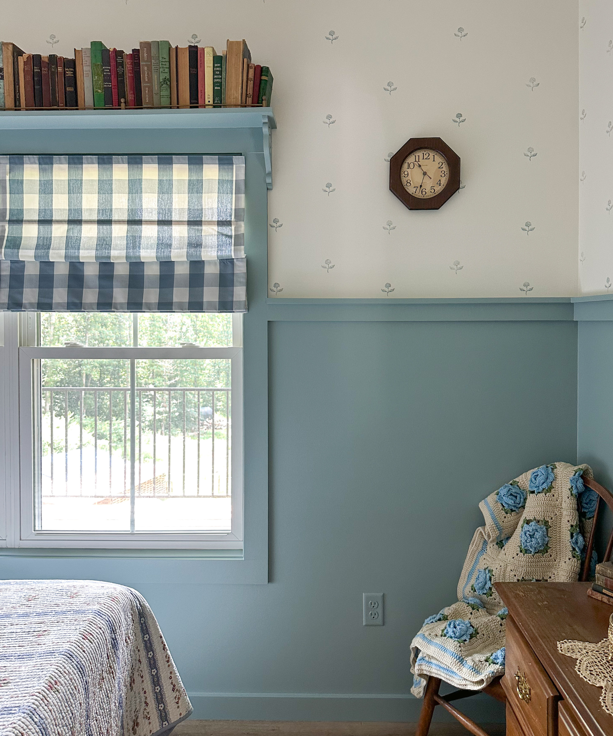 a bedroom with a window covered with gingham shades, a wall split with blue at the bottom and white at the top, a chair with a blanket on it, and a small clock on the wall