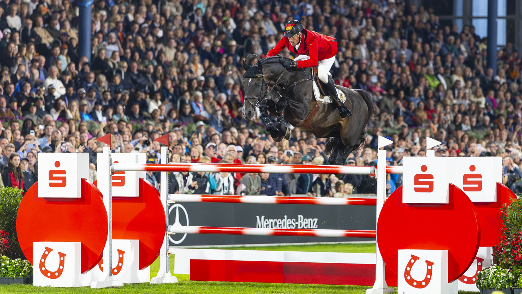 Daniel Deusser, a German rider, and Otello De Guldenboom, a bay horse, jump a red and white fence on the way to helping Germany top the showjumping World Championships team results