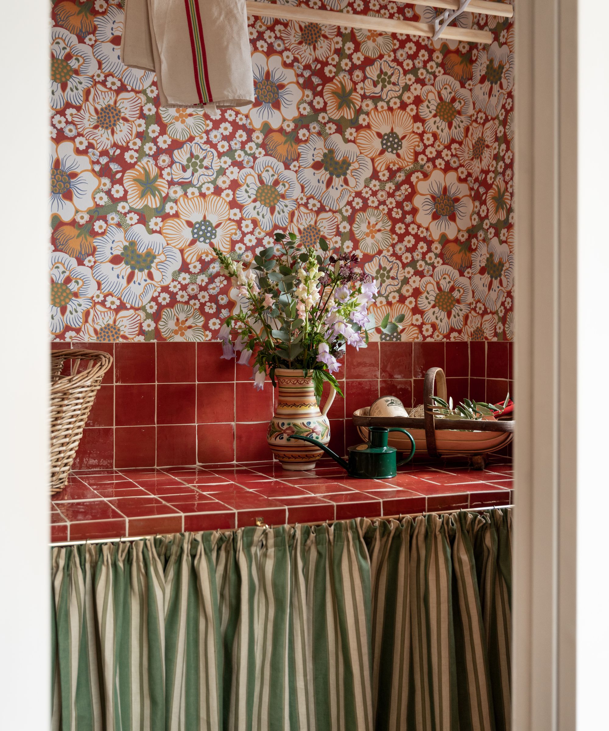 Pantry with floral wallpaper, red tiled countertop and striped cafe curtain hiding storage. A vase of flowers and a tray of produce sit on the counter.