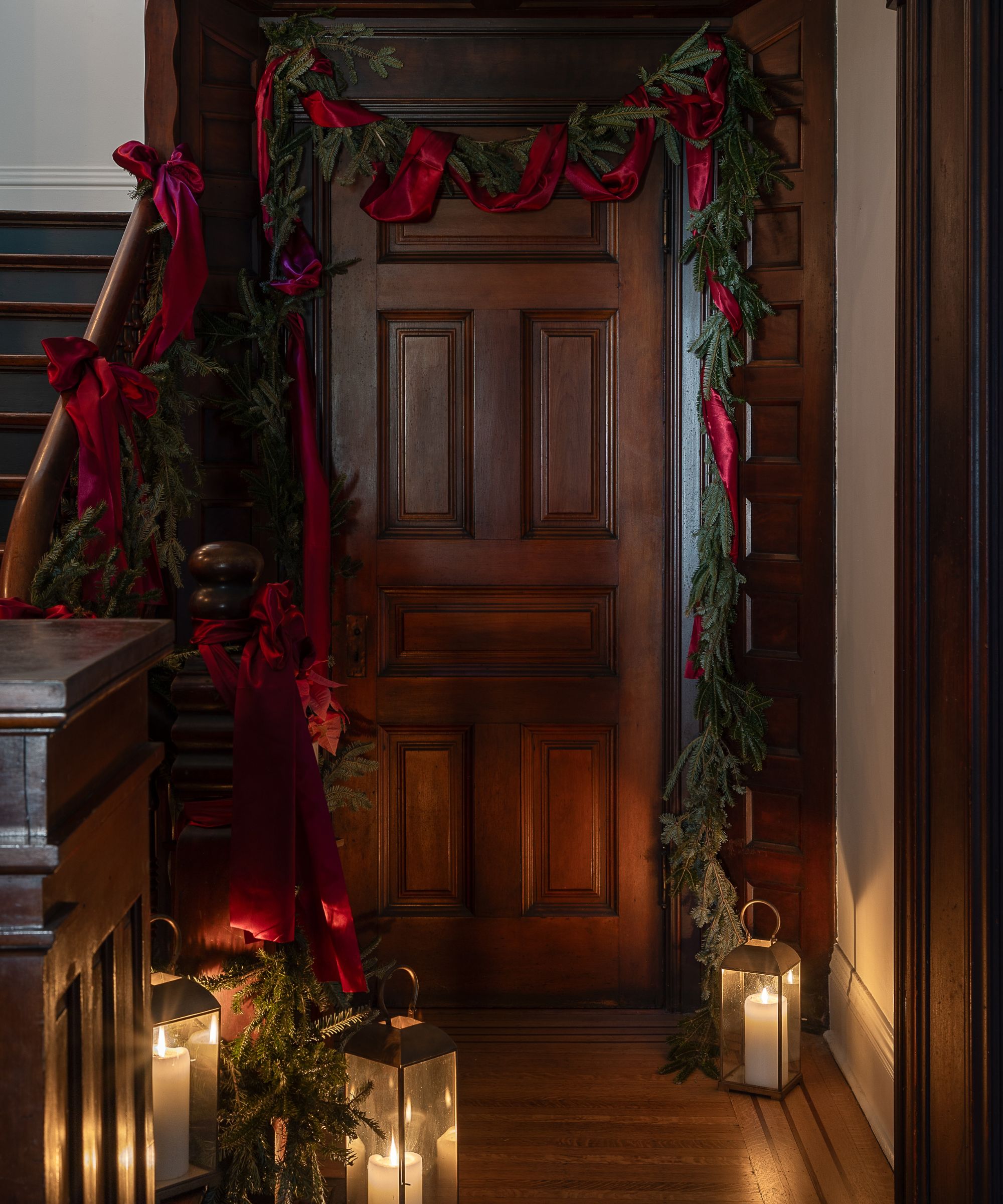 a dark wooden entryway and staircase styled for christmas with fir garlands, red ribbons, and candle lanterns