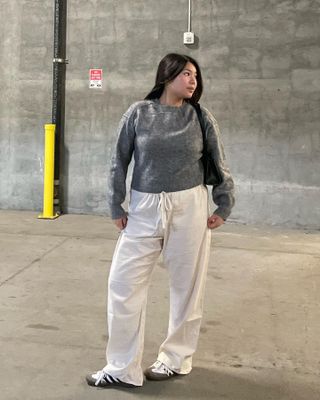 fashion influencer Marina Torres poses in a gray sweater, black shoulder bag, beige linen pants, and white Adidas Samba sneakers