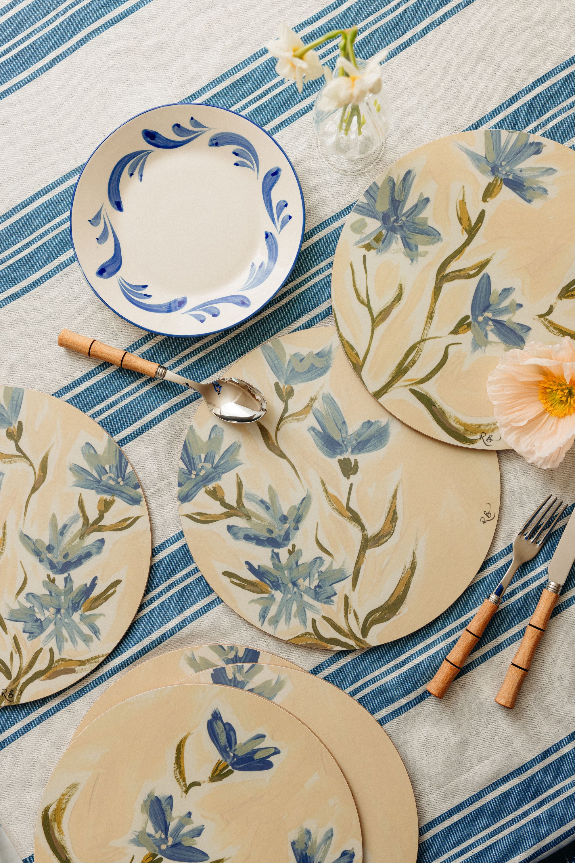 A blue and white striped tablecloth with yellow placemats that are painted with blue and green flowers.