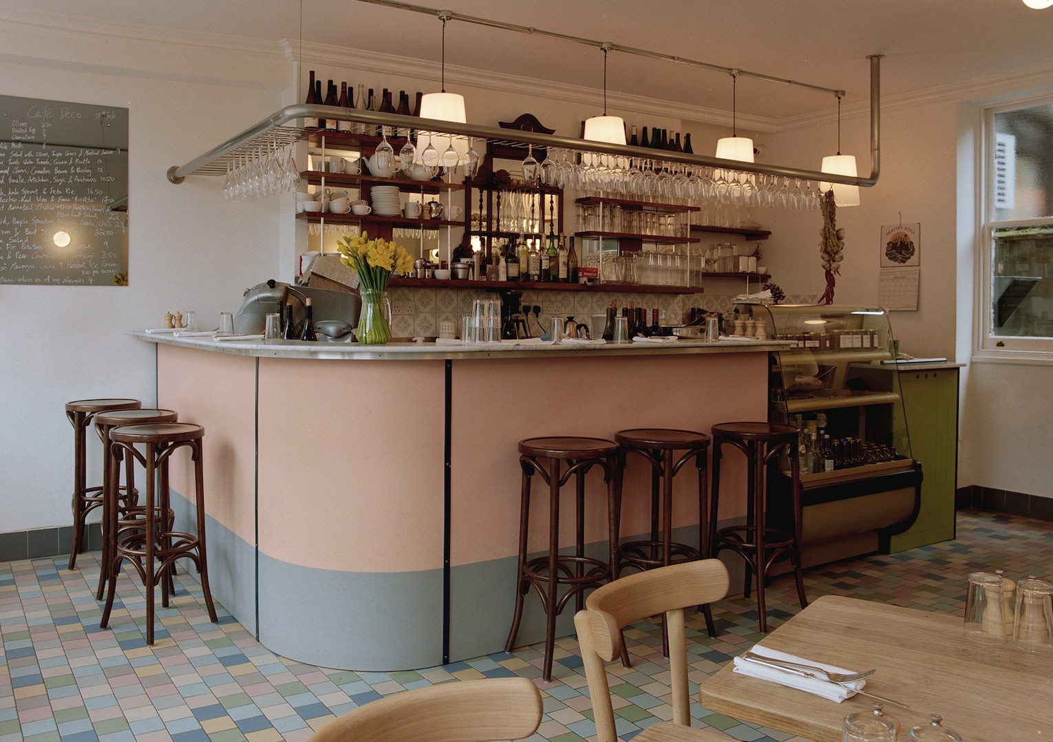 A retro-inspired bistro with checkered, pale pink, green, yellow, white, and blue floors, a pale pink counter, pendant light lamps, wooden tables and chairs, and chrome detailing.