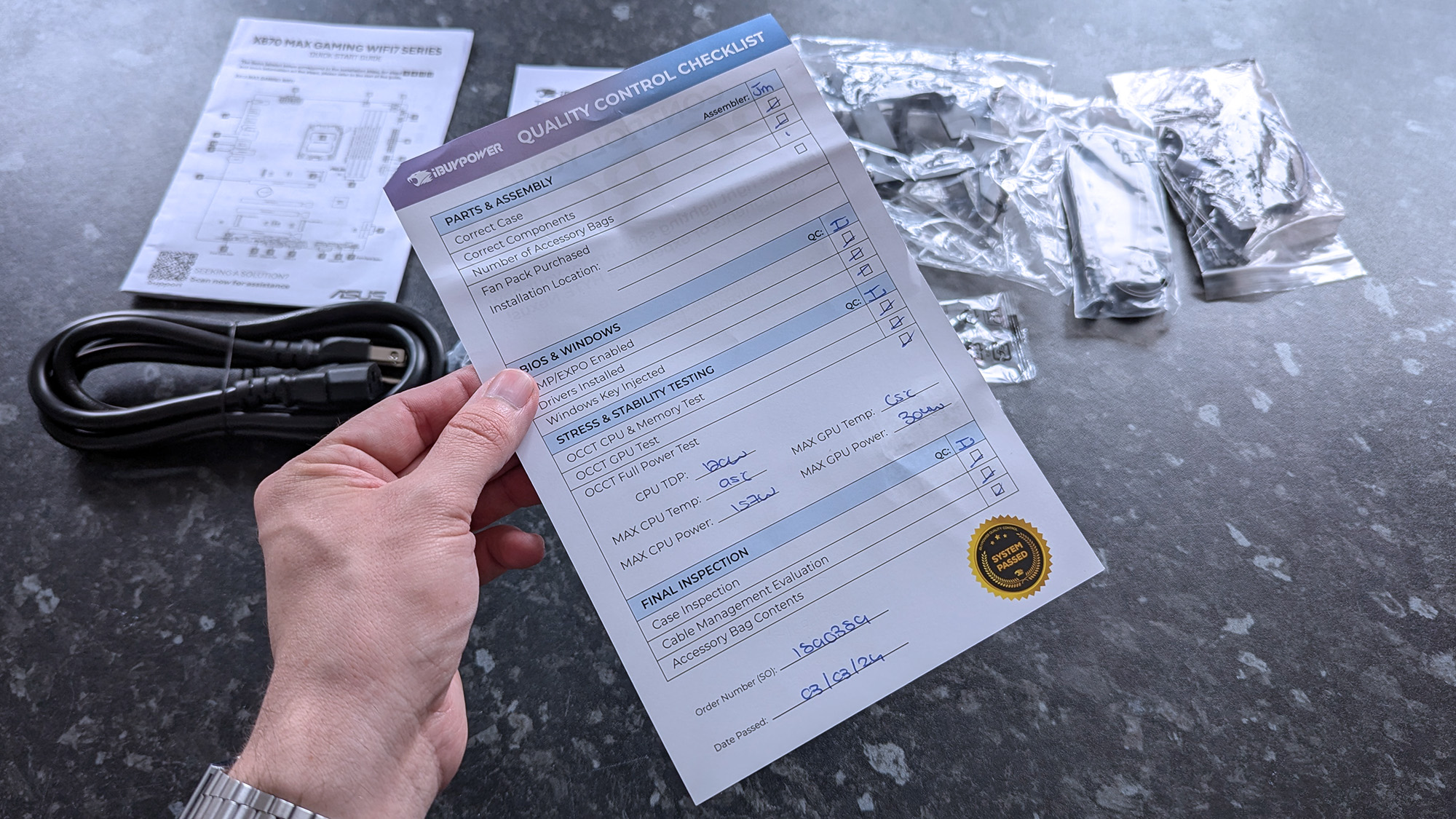 A hand holds a quality control checklist above a dark surface, alongside packaged electronic components, a power cable, and technical sheets.