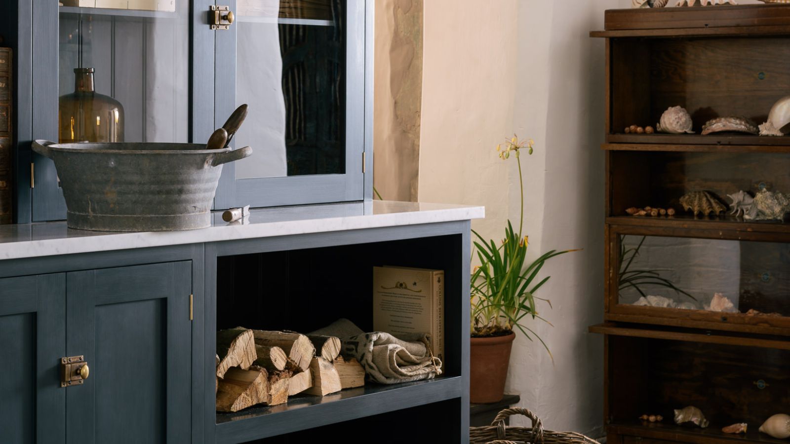 Kitchen utility room showing dark blue cabinet to the left with metal bucket set up for cleaning, logs, cloths and a freestanding shelving cabinet to the right with shells
