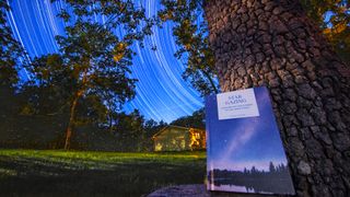 A book leans against a tree against the background of a house in the distance and a dark grassy yard. The night sky is filled with the streaks of stars, traced through the sky through long exposure photos.
