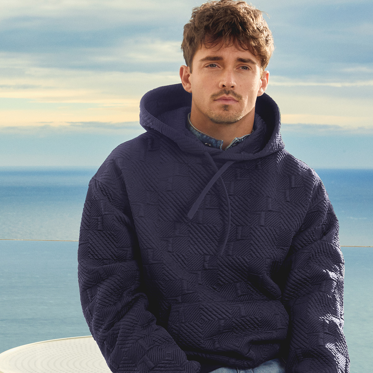 Charles Leclerc wearing a navy-blue hoodie and jeans while posing in front of Monte Carlo.