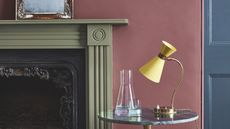Close-up shot of a burgundy-painted wall and a fireplace painted in an olive green trim. There is a small marble side table with a glass carafe and a yellow lamp.
