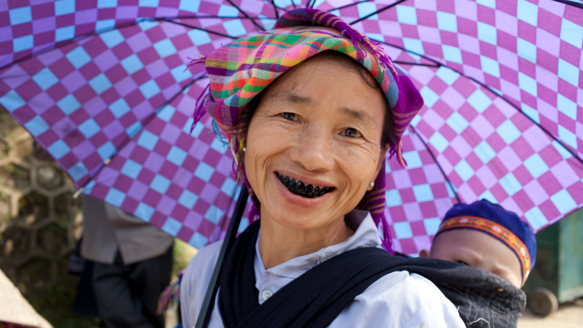 a woman with blackened teeth smiles at the camera; she carries a baby in a pouch on her back and carries a blue and purple checkerboard umbrella