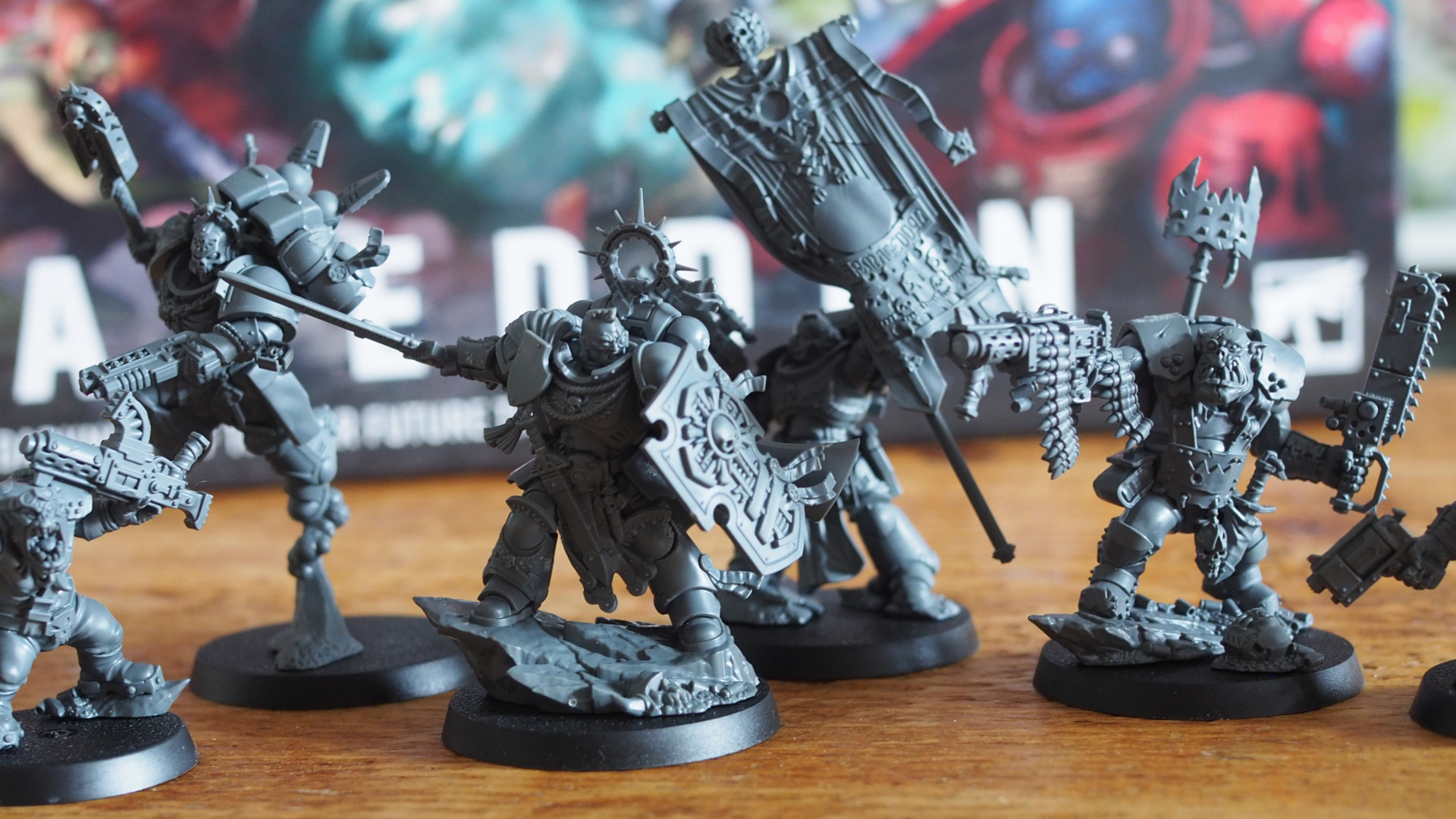 A selection of Space Marine and Ork models in gray plastic arranged on a wooden surface, in front of the Warhammer 40K Armageddon box