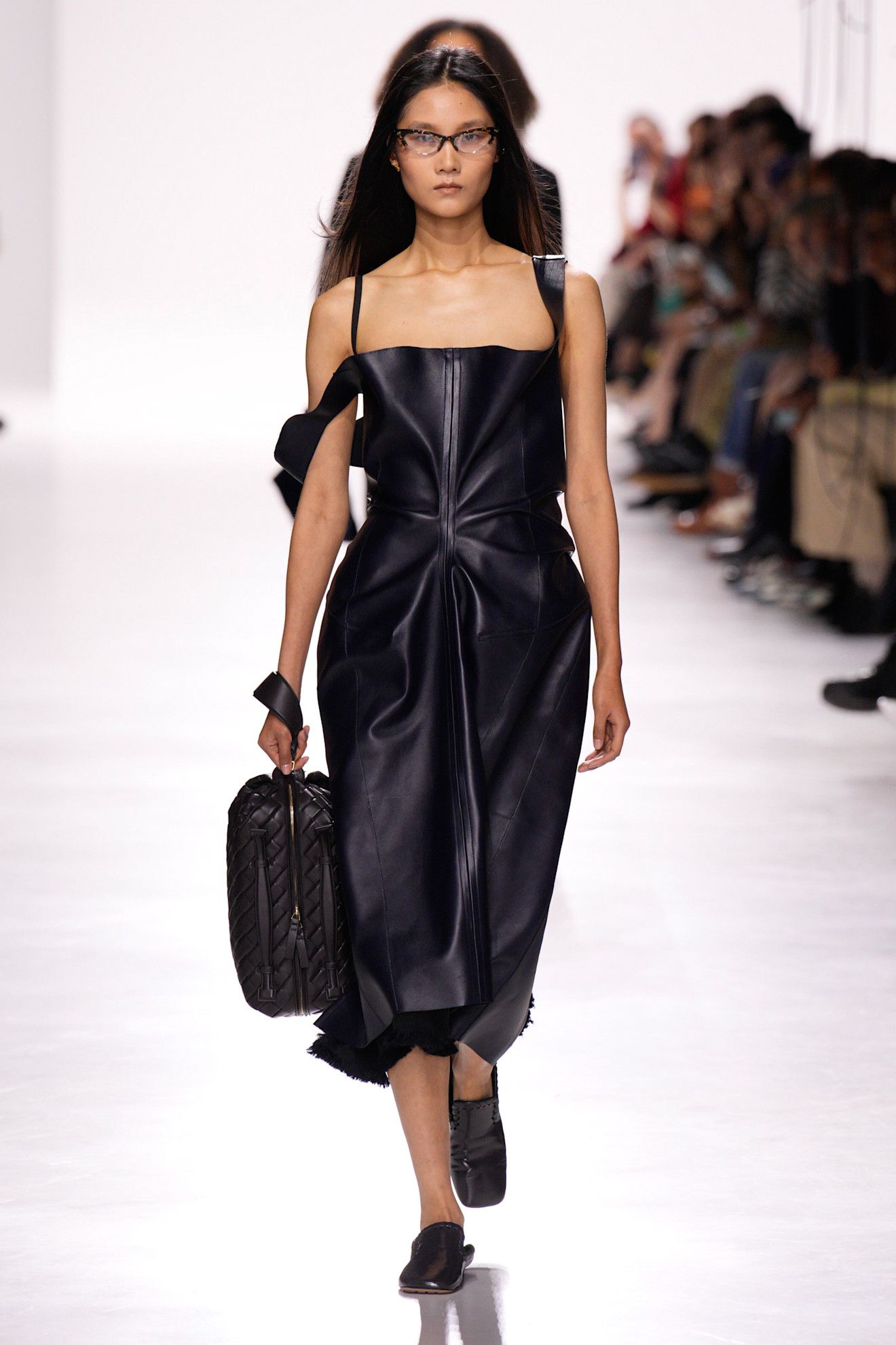 A model at the Bottega Veneta S/S 26 show wearing a leather dress with an Intrecciato dopp kit, slippers, and eyeglasses.