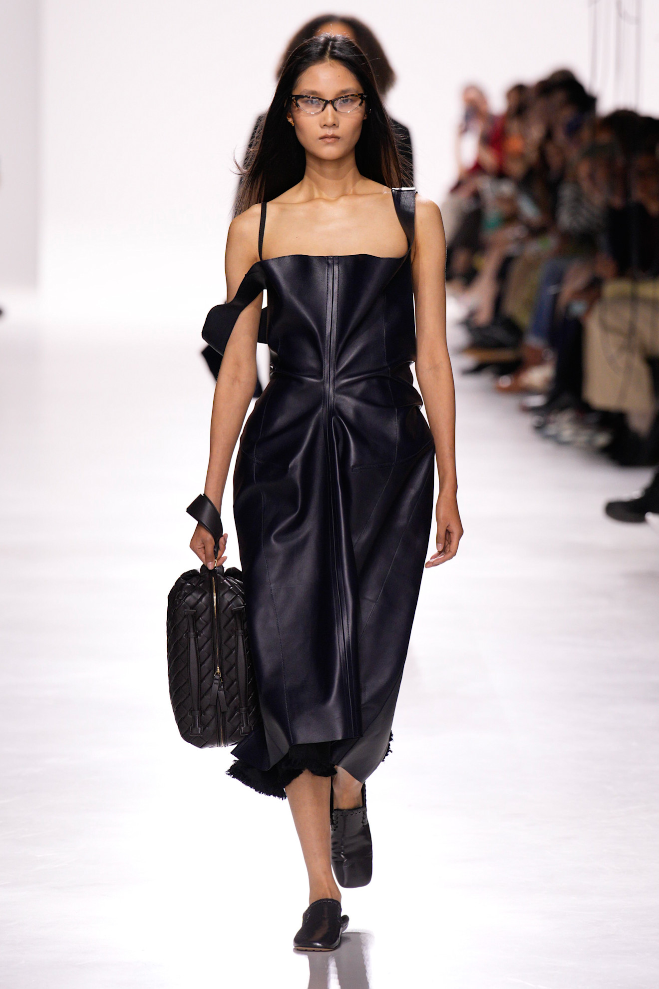 A model at the Bottega Veneta S/S 26 show wearing a leather dress with an Intrecciato dopp kit, slippers, and eyeglasses.