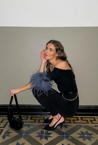 a casual party outfit with a woman in a hallway wearing a black feather top with black leggings, a gold belt, black slingback heels, and a black shoulder bag