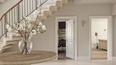 A bright and spacious white entryway with a white staircase with dark banister, white walls, white floors, and a round wooden table with a glass vase of white flowers to the left. To the right are two open doorways with rooms seen beyond.