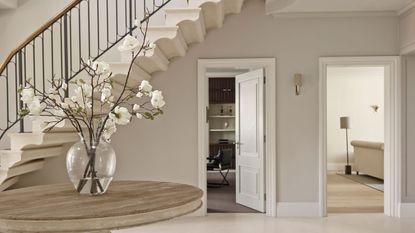 A bright and spacious white entryway with a white staircase with dark banister, white walls, white floors, and a round wooden table with a glass vase of white flowers to the left. To the right are two open doorways with rooms seen beyond.